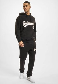 Fekete kapucnis pulóver és melegítőnadrág szett, fehér "Rocawear" felirattal, húzós kapucnival, zsebekkel és puha tapintással. Fehér sportcipő arany részletekkel.