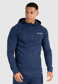Felpa con zip di colore navy, realizzata in tessuto morbido, dotata di cappuccio, maniche lunghe, due tasche con zip e un logo sul petto.