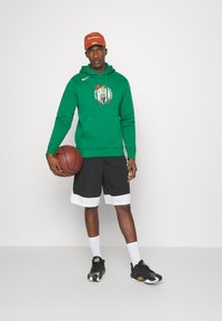 Zelená mikina s logom Boston Celtics, čierne basketbalové šortky s bielym lemom, čierno-zlaté tenisky, drží basketbal. Oranžová čiapka.