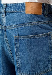 Vaqueros de mezclilla azul con una superficie texturizada, que presentan una etiqueta de cinturón de cuero marrón claro y costuras amarillas en contraste. Incluye un bolsillo trasero.