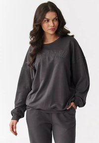 Ciemnoszara bluza o luźnym kroju, z prążkowanymi mankietami i dołem, z napisem "MAKADAMIA" haftowanym na piersi w dopasowanej kolorystyce.