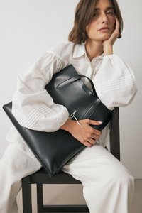 Juodas odinis rankinis krepšys su lygiu paviršiumi ir filigraniniu dizainu. Yra viršutinis rankenas ir dekoratyvinė juostelė. Modelis vilki baltą bliuską.