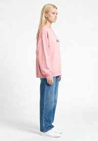Femme blonde en profil portant un sweat rose ample, un jean bleu à jambes larges et des baskets blanches sur un fond blanc uni.