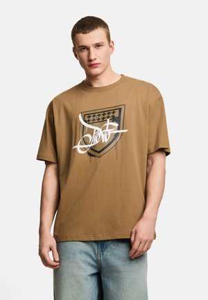 Młody mężczyzna noszący luźną brązową koszulkę z czarno-białym graficznym wzorem tarczy oraz jasnoniebieskie dżinsy, stojący na białym tle.