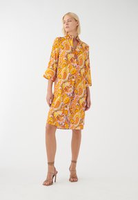 Vestido con estampado de paisley en amarillo y naranja, con cuello y mangas tres cuartos, hecho de una tela ligera. La modelo lleva tacones nude con tiras.