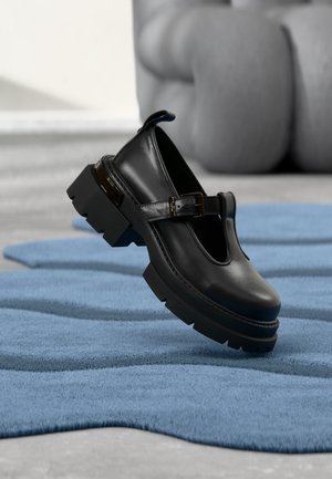 Chaussure Mary Jane en cuir noir avec semelle épaisse et bride à boucle, présentée flottant au-dessus d'un tapis texturé bleu sur un sol gris.