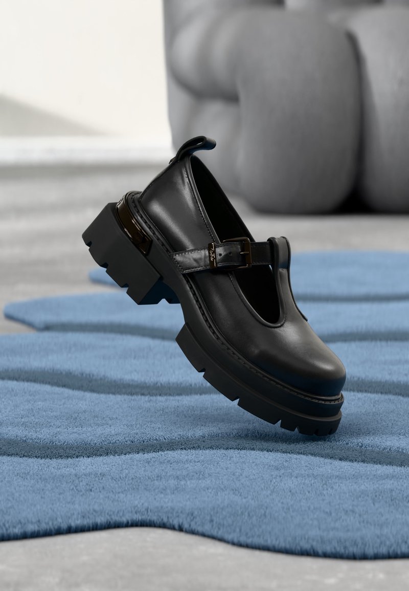 Chaussure Mary Jane en cuir noir avec semelle épaisse et bride à boucle, présentée flottant au-dessus d'un tapis texturé bleu sur un sol gris.