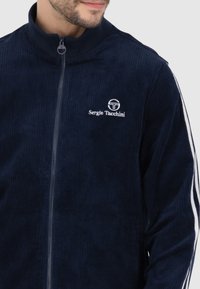 Marineblauwe corduroy zip-up jas met witte zijstrepen en het "Sergio Tacchini" logo in witte borduursel op de borst.