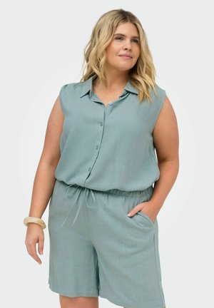 Žena v tielkovom modrozelenom topánku s gombíkmi a zodpovedajúcich šortkách so šnúrkou, stojaca s jednou rukou v vrecku a neutrálnym pozadím.