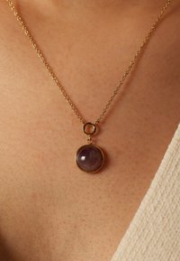 Collier en or avec un design en maillons, mettant en avant un pendentif en forme de pierre précieuse violette, fixé dans un cadre circulaire. Texture lisse, style minimaliste.