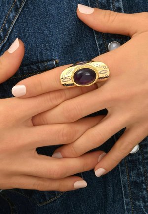 Bague en or avec une pierre ovale violet, présentant des détails et un design complexes. Portée sur une main aux ongles clairs contre un tissu en denim.