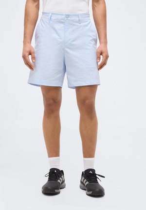 Shorts bleu clair arrivant aux genoux portés avec des chaussettes blanches et des baskets Adidas noires, sur une personne debout devant un fond uni.