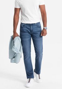 Vaqueros de mezclilla azul oscuro con corte recto, combinados con una camisa de botones azul claro apoyada sobre un brazo y zapatillas blancas con acentos a rayas.