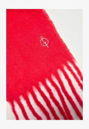 Jaskrawoczerwona wełniana szalik z frędzlami na końcach i małym białym wyhaftowanym logo blisko jednego brzegu na jasnym tle.