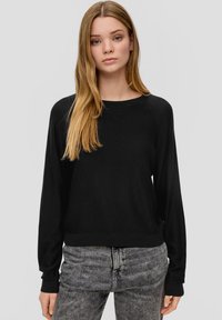 Schwarzes Langarmoberteil mit Rundhalsausschnitt und geripptem Saum, kombiniert mit grauen Jeans. Der Stoff wirkt glatt und figurbetont.