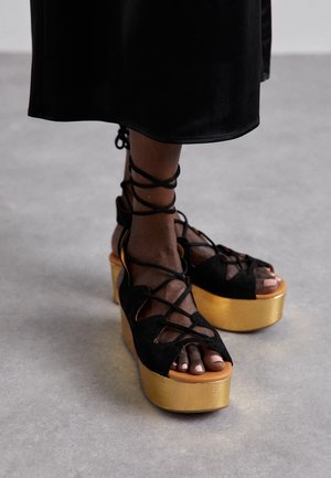 Pieds en sandales noires à lacets avec talons compensés dorés sur un sol gris, portant une jupe noire.