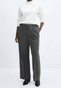Personne portant un haut blanc à manches longues, un pantalon gris foncé à cordon de serrage et des bottines noires, debout devant un fond clair uni.