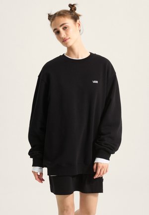 Černá oversized mikina s manžetovými rukávy, která má malé bílé logo Vans. Nositelná přes černou sukni, ukazující uvolněný střih.