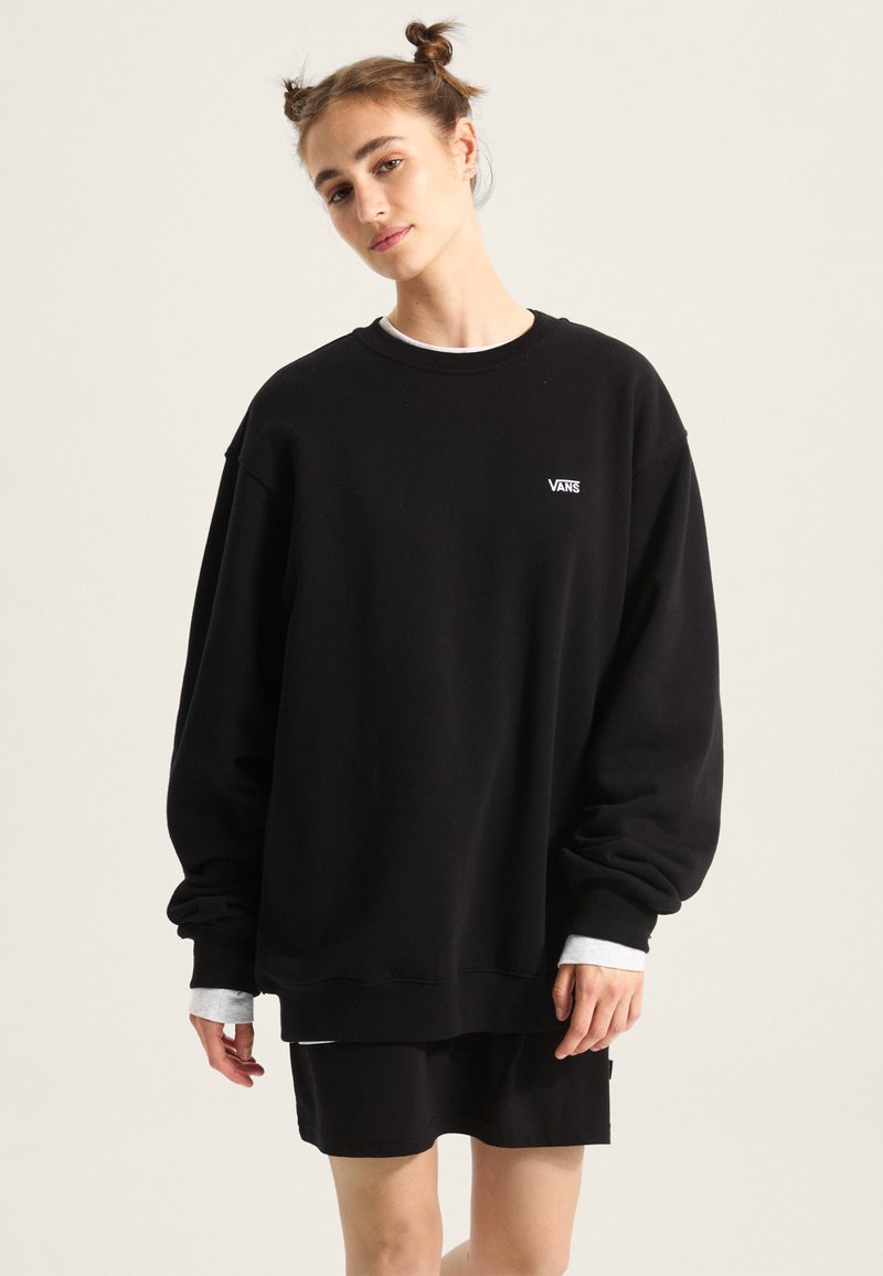 Black oversized sweatshirt with cuffed sleeves, featuring a small white Vans logo. Worn over a black skirt, showcasing a relaxed fit.