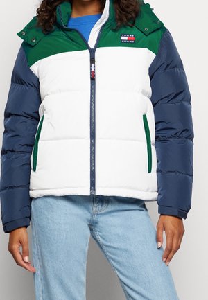 Kvinde iført en farveblokeret dunjakke i grøn, hvid og marineblå med lynlås foran og lyseblå jeans.