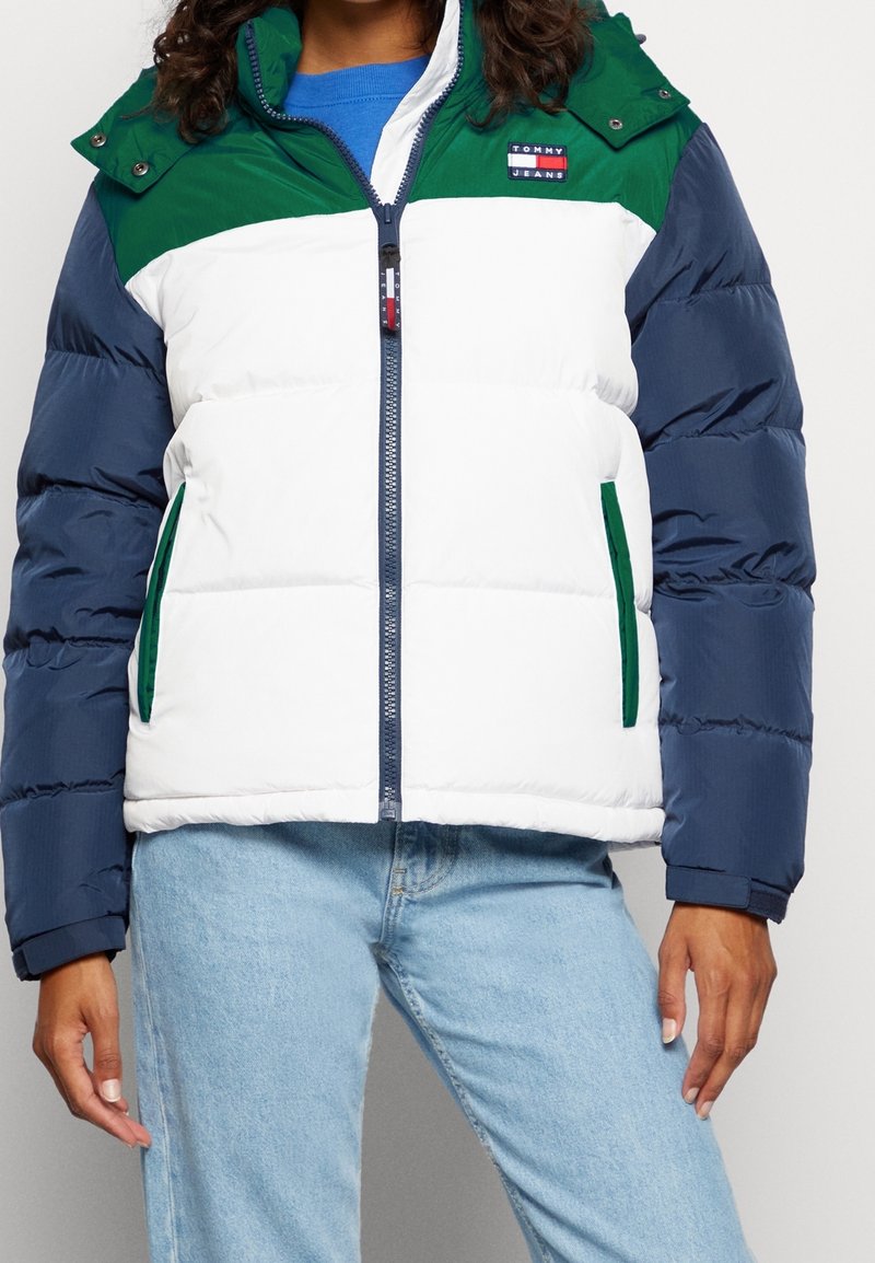Femme portant une doudoune color block verte, blanche et bleu marine avec une fermeture éclair devant et un jean bleu clair.