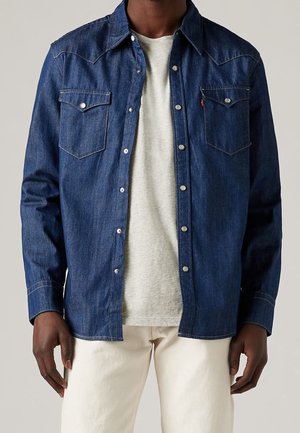 Camisa de mezclilla en azul oscuro con un diseño estructurado, botón a presión, dos bolsillos en el pecho y detalles de costura en contraste. Combinada con una camiseta gris claro.