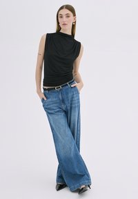 Haut sans manches noir avec design drapé, associé à un jean en denim bleu à jambes larges et des escarpins noirs à bout pointu, présenté sur un fond clair.