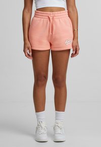 Peachfärgade shorts i bomullsblandning med elastisk midja, sidofickor och broderad logotyp; kombineras med vita sneakers och ankelsockor.