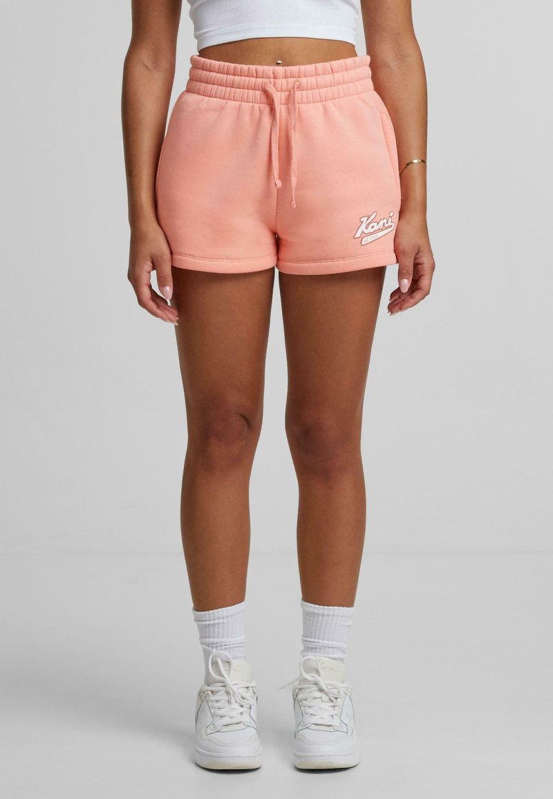 Peachfärgade shorts i bomullsblandning med elastisk midja, sidofickor och broderad logotyp; kombineras med vita sneakers och ankelsockor.