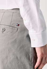 Lysegrå bukser med en glatt tekstur har en knappelukking og en liten brodert logo i marineblå, rød og hvit. En hvit skjorteerme er synlig.
