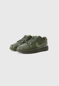 Žalios Nike sportbačiai su tekstūruotu viršumi, turinčiu krokodilo odos raštą, apvaliu nosiu ir plokščiu guminiu padu. Šnervės atitinkamos žalios spalvos.
