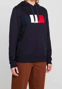 Námořnická mikina s barevně rozdělenou hrudní částí, která obsahuje červené, bílé a modré sekce. Vyrobena z měkké tkaniny, s nastavovací šňůrkou a žebrovanými manžetami.