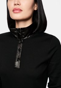 Schwarzes Oberteil mit einem hohen, glänzenden schwarzen Kragen und einem geknöpften Verschluss. Weicher Stoff mit glatter Textur.