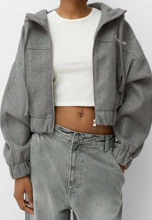 Person trägt einen kurzen grauen Kapuzenpullover mit Reißverschluss, weißes kurzes Top, lockere graue Hose und einen Bauchnabelpiercing, mit der Hand in der Tasche.