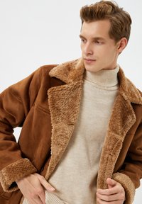 Ung mand justerer sin brune shearlingjakke over en beigefarvet striktrøje, mens han ser let til siden mod en ensfarvet baggrund.