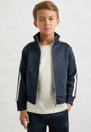 Tmavomodrá bunda na zips s bielymi pruhmi po stranách, rebrovanými manžetami a golierom. Nosená cez krémovú košeľu. Na prednej strane je logo. Hladká látka, ležérny strih.