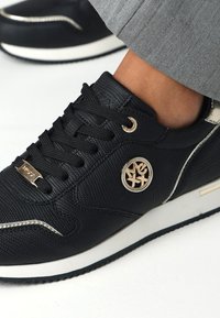 Czarne trampki o teksturowanym wykończeniu, z sznurówkami, złotym emblematem, białymi akcentami i czarną gumową podeszwą. Projekt cechuje niski profil.