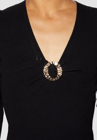 Nahaufnahme einer Person, die ein schwarzes, geripptes Strickoberteil mit einem tiefen V-Ausschnitt und einem dekorativen, glänzenden goldenen Ring in der Mitte trägt.