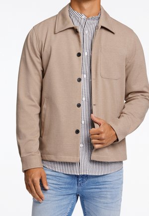 Mężczyzna ubrany w beżową zapinaną na guziki kurtkę na białą i szarą koszulę w paski, w jasnoniebieskich dżinsach, trzymający klapę kurtki prawą ręką.