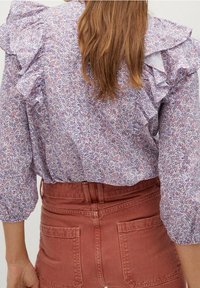 Bluse mit floralem Muster und Rüschensäumen in Lila und Pink auf hellem Hintergrund; kombiniert mit hochgeschnittenen rostfarbenen Hosen.