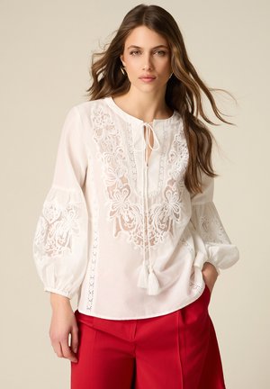 Kobieta ubrana w białą haftowaną bluzkę z frędzlami i obszernymi rękawami, w połączeniu z czerwonymi spodniami, stojąca na beżowym tle.