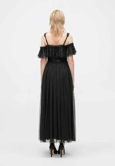 Robe de soirée noire avec des manches en tulle dénudées, un corsage orné et une jupe plissée fluide. Portée avec des talons hauts noirs.