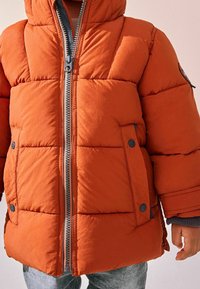 Veste orange matelassée avec capuche, dotée d'une fermeture éclair frontale, de deux poches à boutons-pression et d'un tissu texturé avec des coutures diagonales.