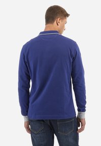 Långärmad pikétröja i marinblå bomull med grå manschettaccent. Har en klassisk krage och avslappnad passform, med en ren baksidestjänst.