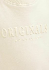 Krämfärgad sweatshirt med präglad logotext "ORIGINALS JACK & JONES" på ett texturerat tyg. Inkluderar en ribbad halsringning.