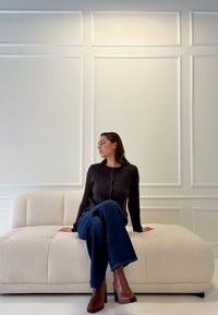 Lenkta kreminė sofa su purus faktūra ir siuvinėtais mygtukais, derinama su rudais megztais kardiganais ir mėlynais platėjančiais džinsais.