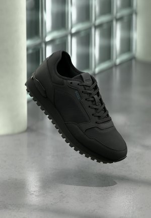 Czarny sneaker marki BOSS z fakturowaną tkaniną i skórzanymi panelami, zawiązanymi sznurówkami, unoszący się nad szarą betonową podłogą w pobliżu szklanych bloków.