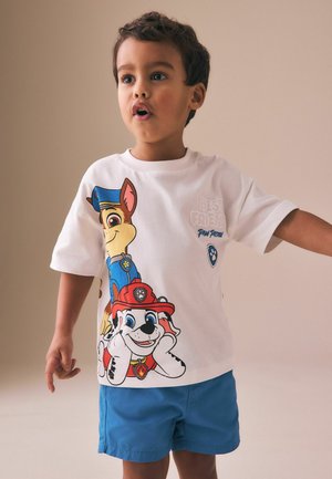 Junger Junge mit braunen Haaren, der ein weißes Paw Patrol T-Shirt und blaue Shorts trägt, steht mit leicht ausgestreckten Armen da, der Mund geöffnet.