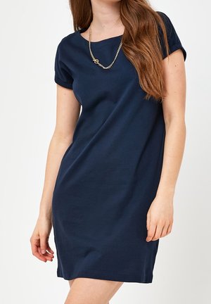 Kobieta ubrana w granatową sukienkę z krótkim rękawem i złoty łańcuszek na jednolitym białym tle.