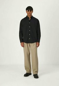 Chemise noire à boutons, pantalon beige à jambes larges, chaussures noires ; modèle portant des lunettes de soleil, cheveux courts. Fond uni.